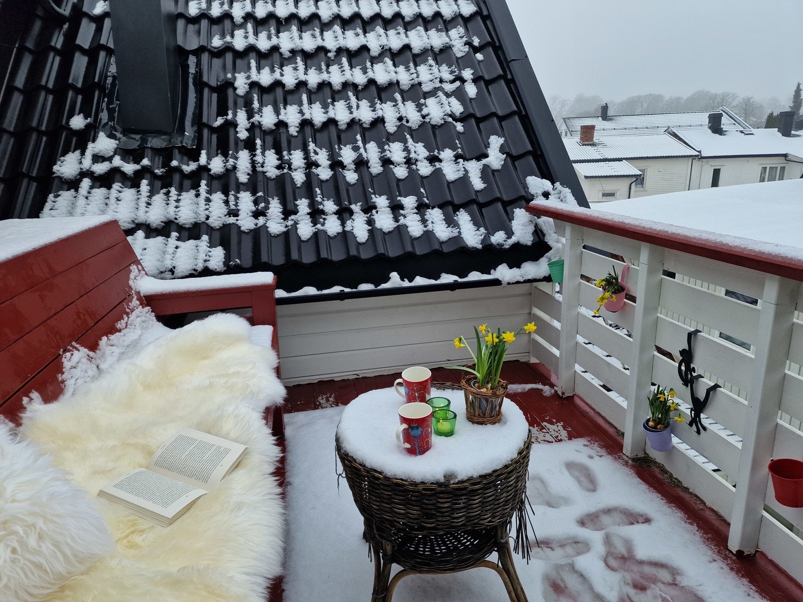 En koselig balkong med rødt gulv og rekkverk. Utsikt mot nærliggende hus og snødekte tak Galleribilde