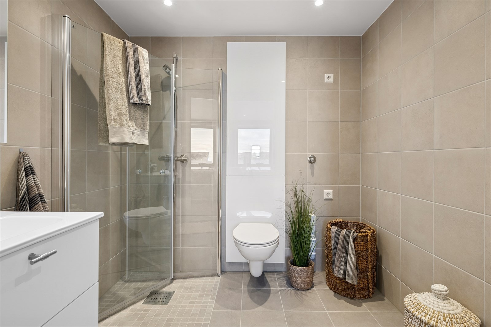 Dusjhjørnet har innfellbare glassdører og veggmontert dusjsett med hånddusj. Videre inneholder badet vegghengt toalett og opplegg for vaskemaskin. Galleribilde
