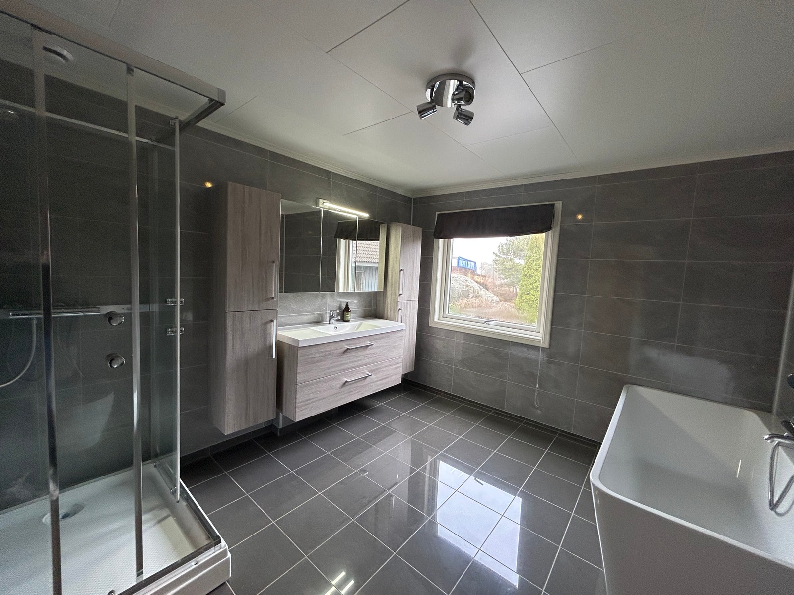 Nytt moderne bad med fliser på gulv og veggplater med flisemønster. Vinduet gjør at du får godt med lys inn på badet. Toalett, badekar, dusjkabinett og vegghengt servant med plass til mye oppbevaring. Galleribilde