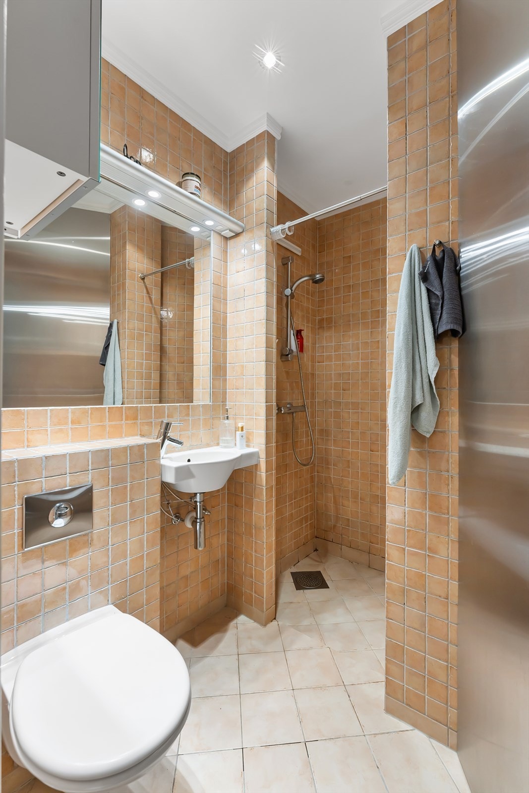 Bad 2 med nedsenket dusjhjørne, vegghengt servant, speilfelt innmurt i flis over servant, vegghengt toalett og flislagt gulv med varmekabler. Galleribilde