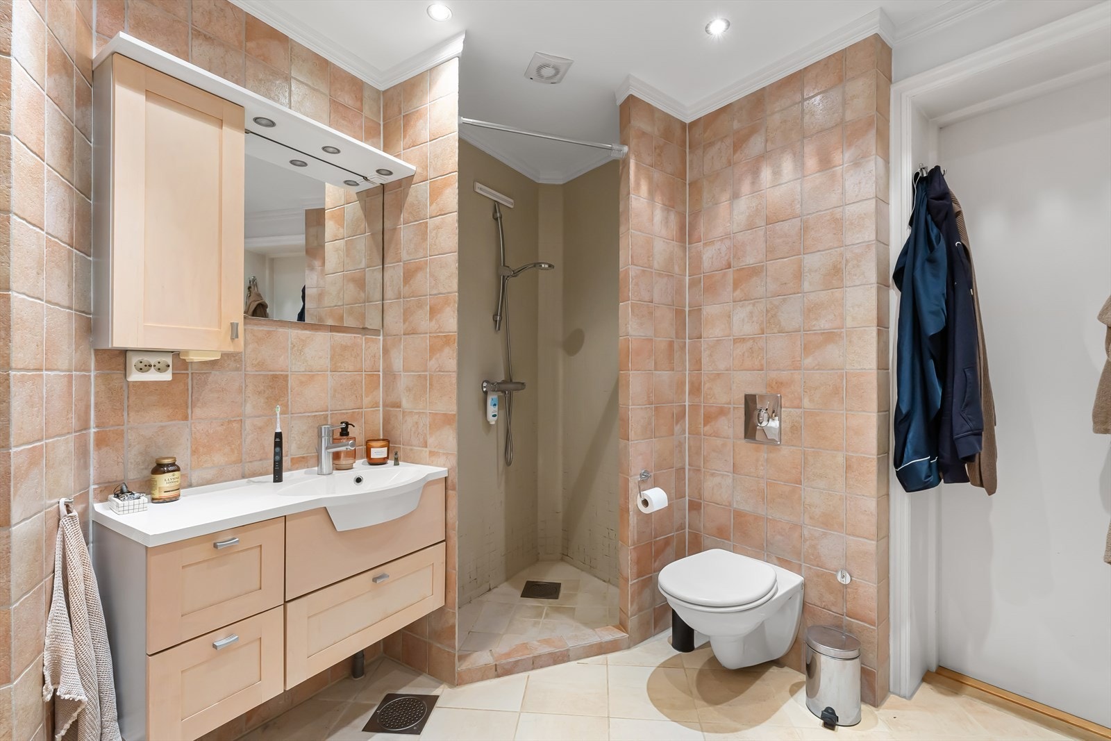 Bad med innmurt flislagt badekar og nedsenket dusjhjørne, helservant med underskuffer, skap, speil og downlights over servant, veggmontert høyskap, speilfelt innmurt i flis på vegg ved badekar og vegghengt toalett. Galleribilde