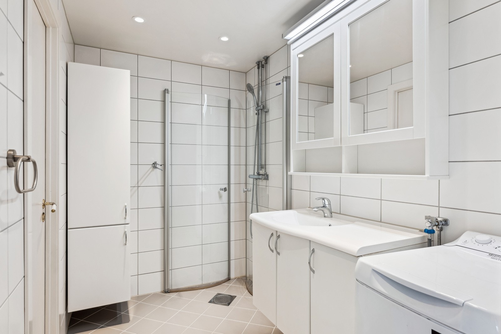 Baderomsinnredning med servant med underskap, veggskap og speilfronter med overlys. Dusj med inn-/utadslående glassvegger og det er opplegg for vaskemaskin. Galleribilde