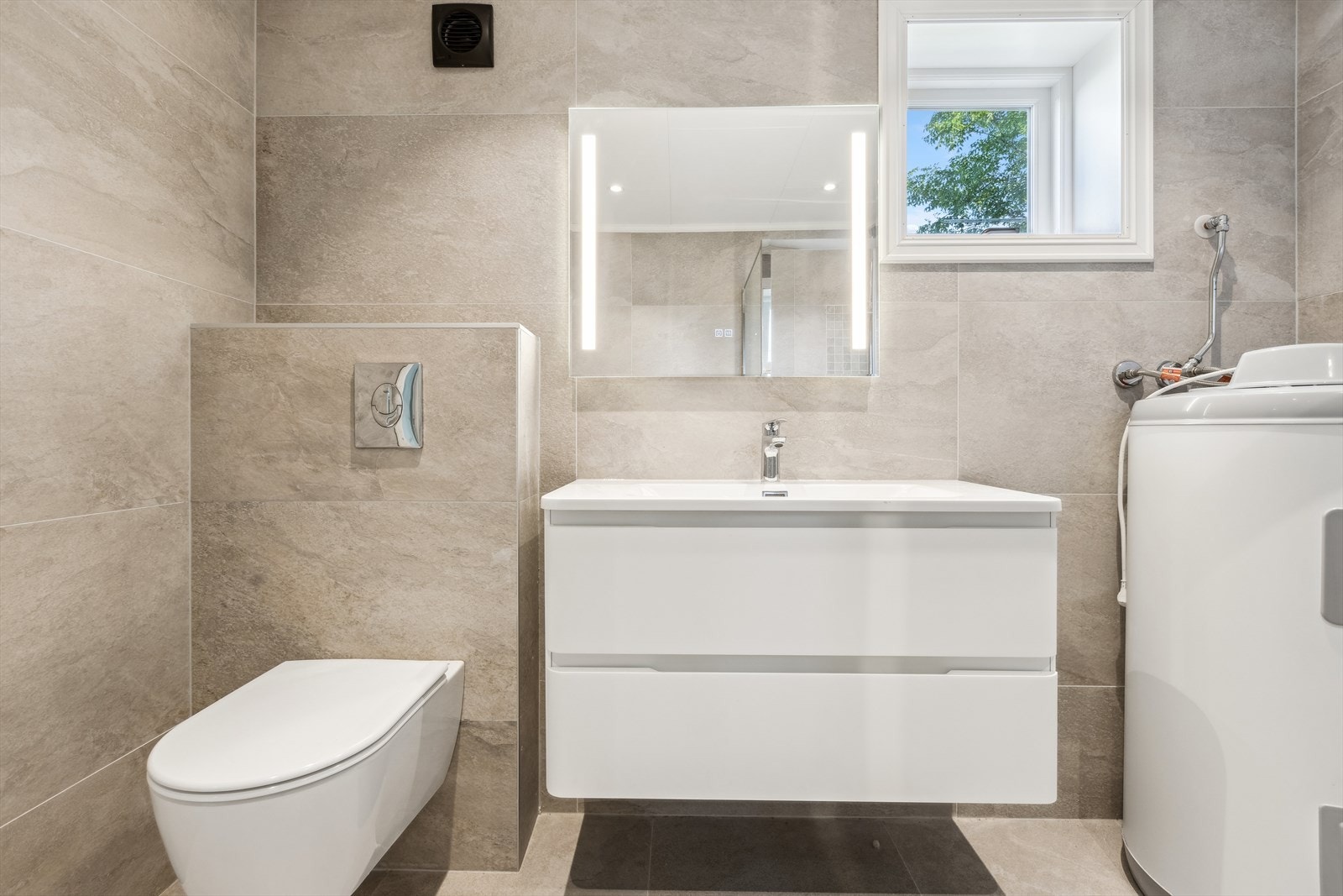 Videre er rommet innredet med baderomsinnredning med nedfelt servant, speil, vegghengt toalett og dusjhjørne. Videre er det varmtvannsbereder montert her og det er opplegg for vaskemaskin her. Galleribilde