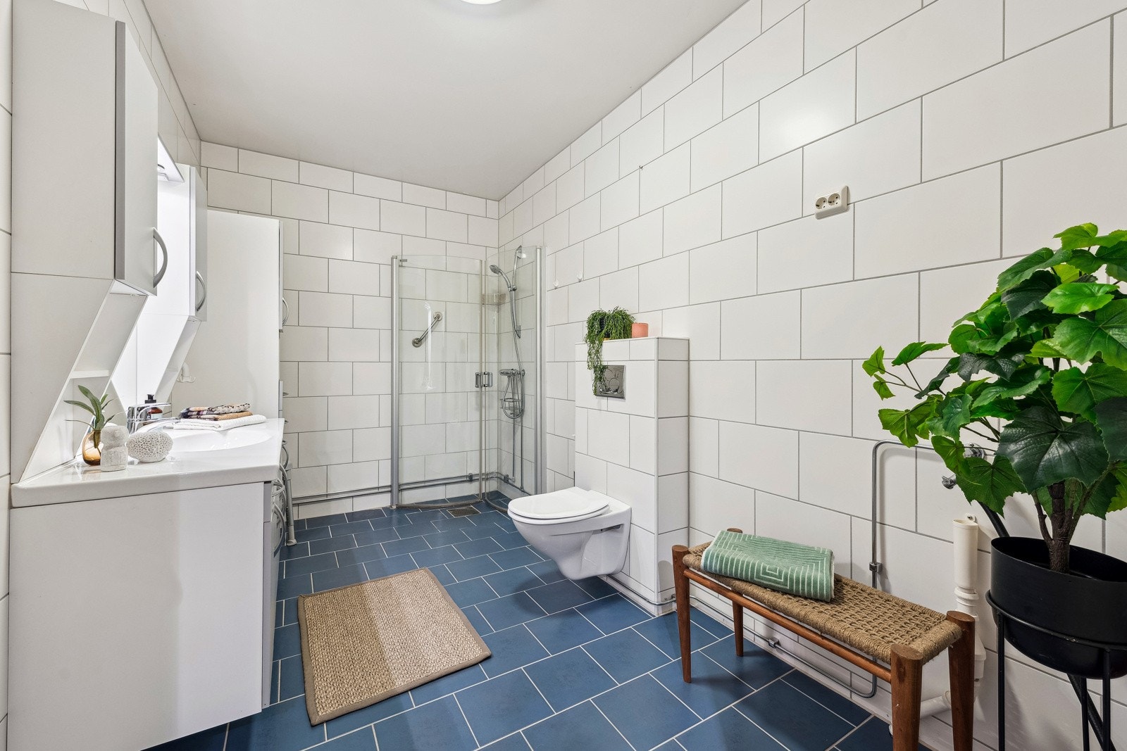 Romslig baderom som er utstyrt med dusj med inn-/utadslående dusjvegger, servant med underskap, speil med overlys, veggskap og veggklosett. Galleribilde