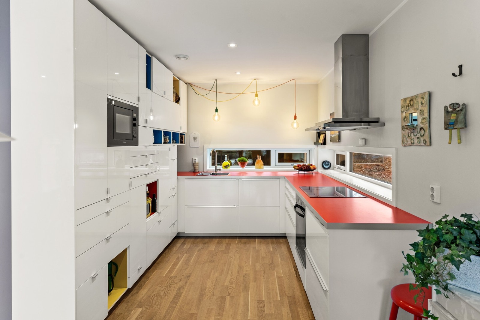 Den hvite kjøkkeninnredningen har en rød benkeplate av laminat og fargerike detaljer i utvalgte hyller, noe som tilfører liv og dynamikk til rommet. Galleribilde