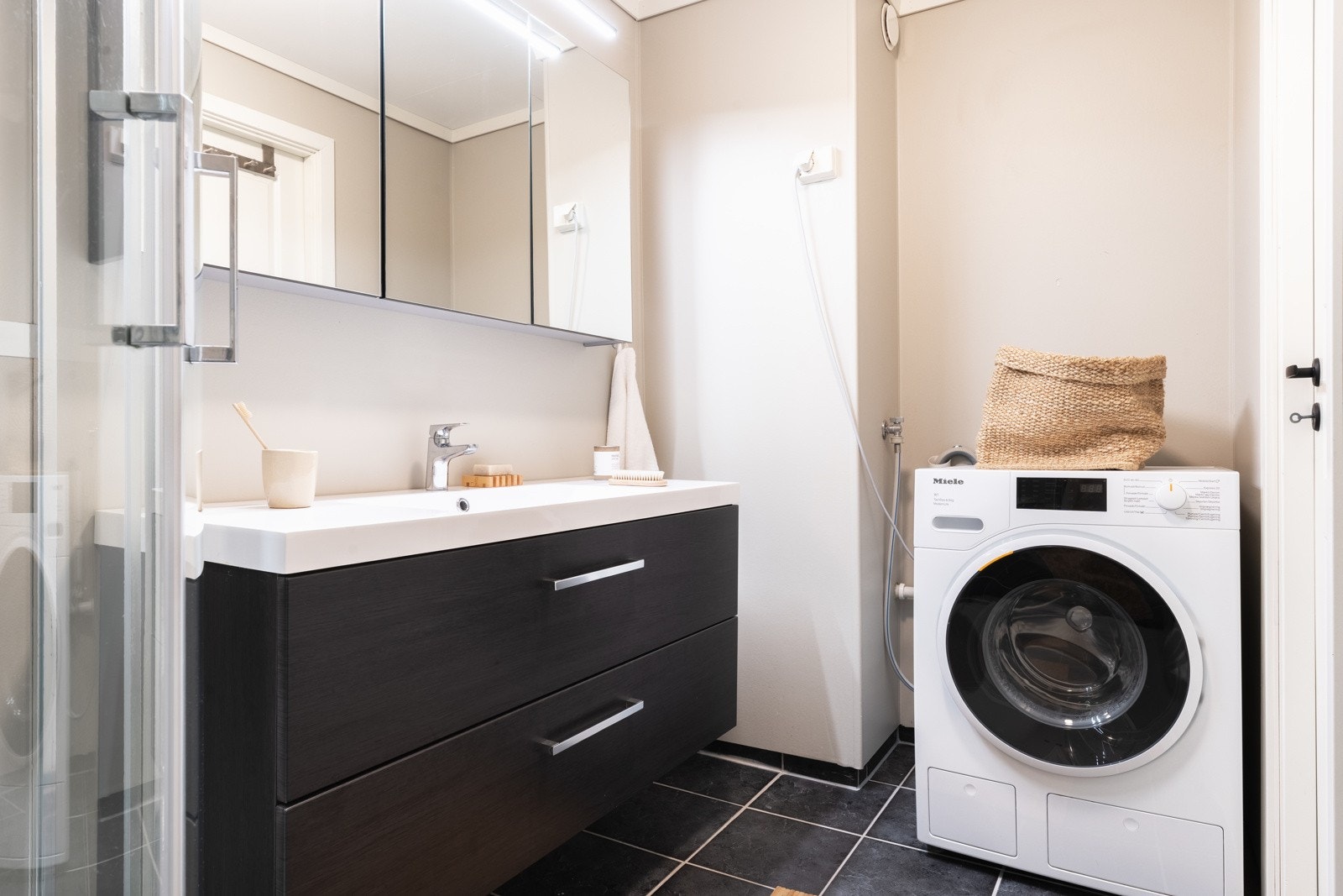 Et tidløst bad i moderne fargetoner har tilkomst fra kjøkkenet og måler 3,8 kvm. Badet har fliser med varmekabler på gulvet og malte baderomsplater på veggene. Galleribilde
