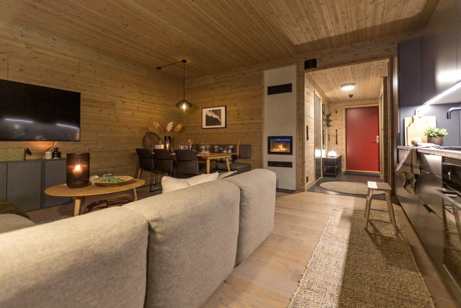 Stue med lys parkett og panel på vegger og tak. Peis og åpen kjøkkenløsning. Galleribilde