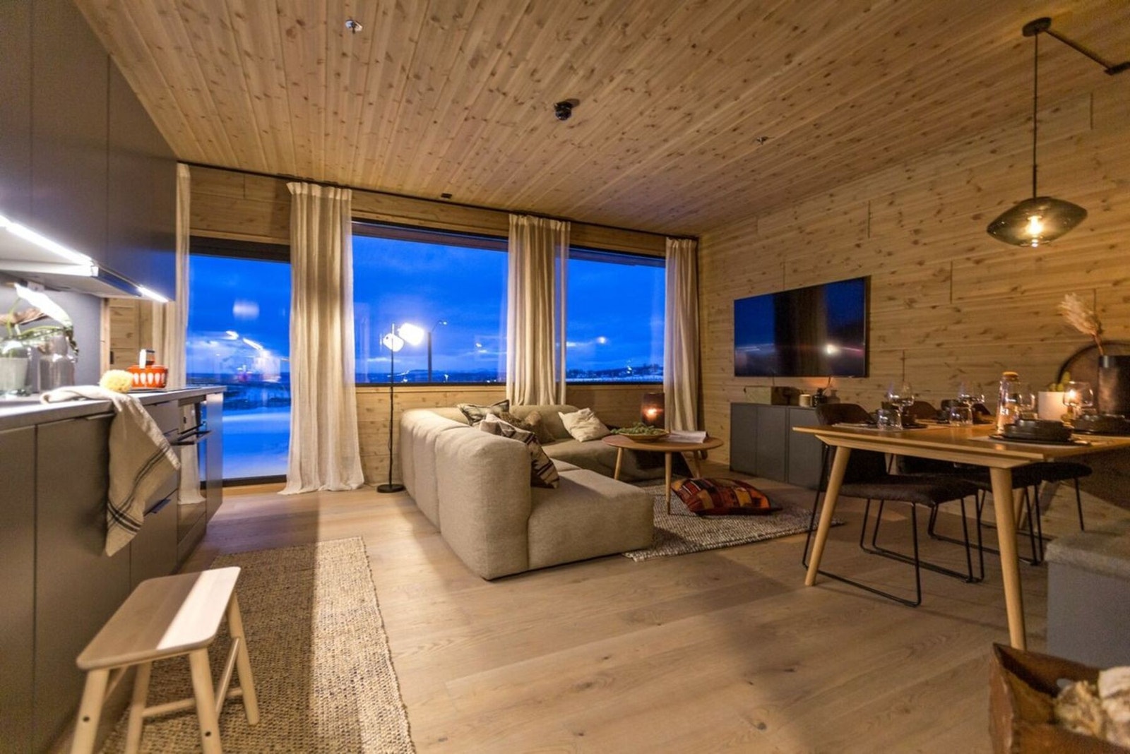 Stue med åpen kjøkkenløsning, store vinduer som gir mye lys og utsikt. Tregulv og trepanel på vegger og tak gir en varm atmosfære. Rommet har en spiseplass og sofakrok. (Avvik på utsikt fra slik denne leiligheten fremstår) Galleribilde