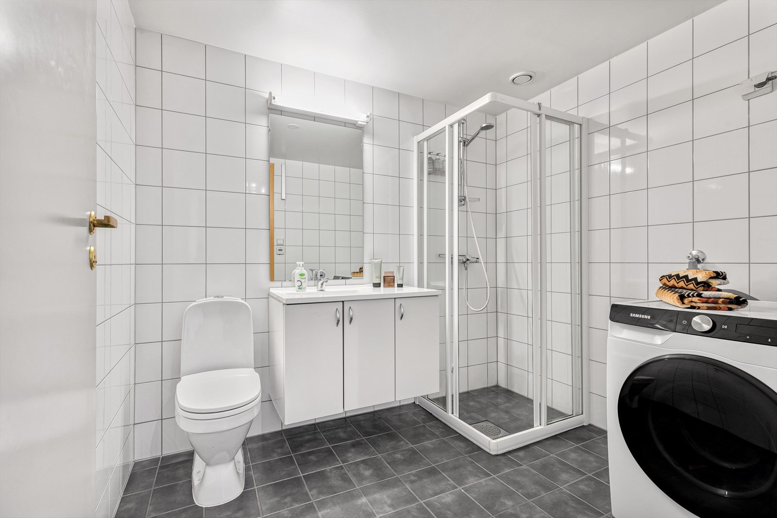 Badet er utstyrt med servantinnredning med underskap, speil med belysning over, gulvstående toalett og dusjhjørne med skyvedører i glass. Det er opplegg for og plass til vaskemaskin. Galleribilde