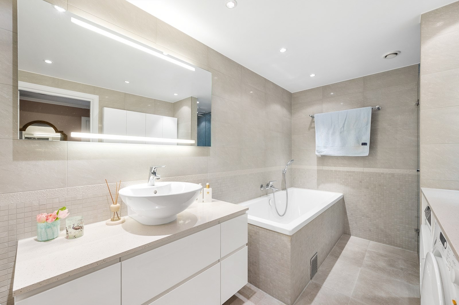 Moderne baderomsinnredning med benkeplate i kompositt, servant og lysspeil. Badekar med armatur og hånddusj Galleribilde