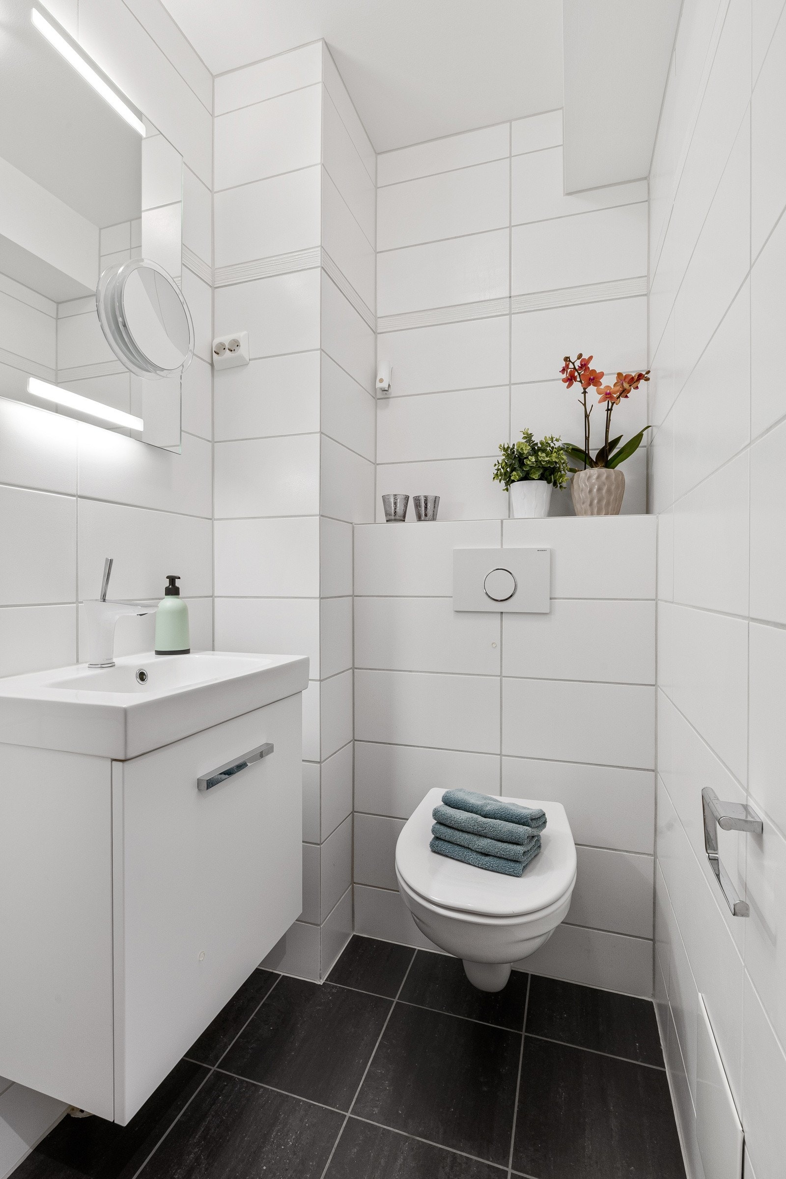 Toalettrommet er moderne og stilrent, med flislagte vegger og gulv. Det er innredet med en servant med underskap, speil med belysning og et veggmontert toalett. Galleribilde