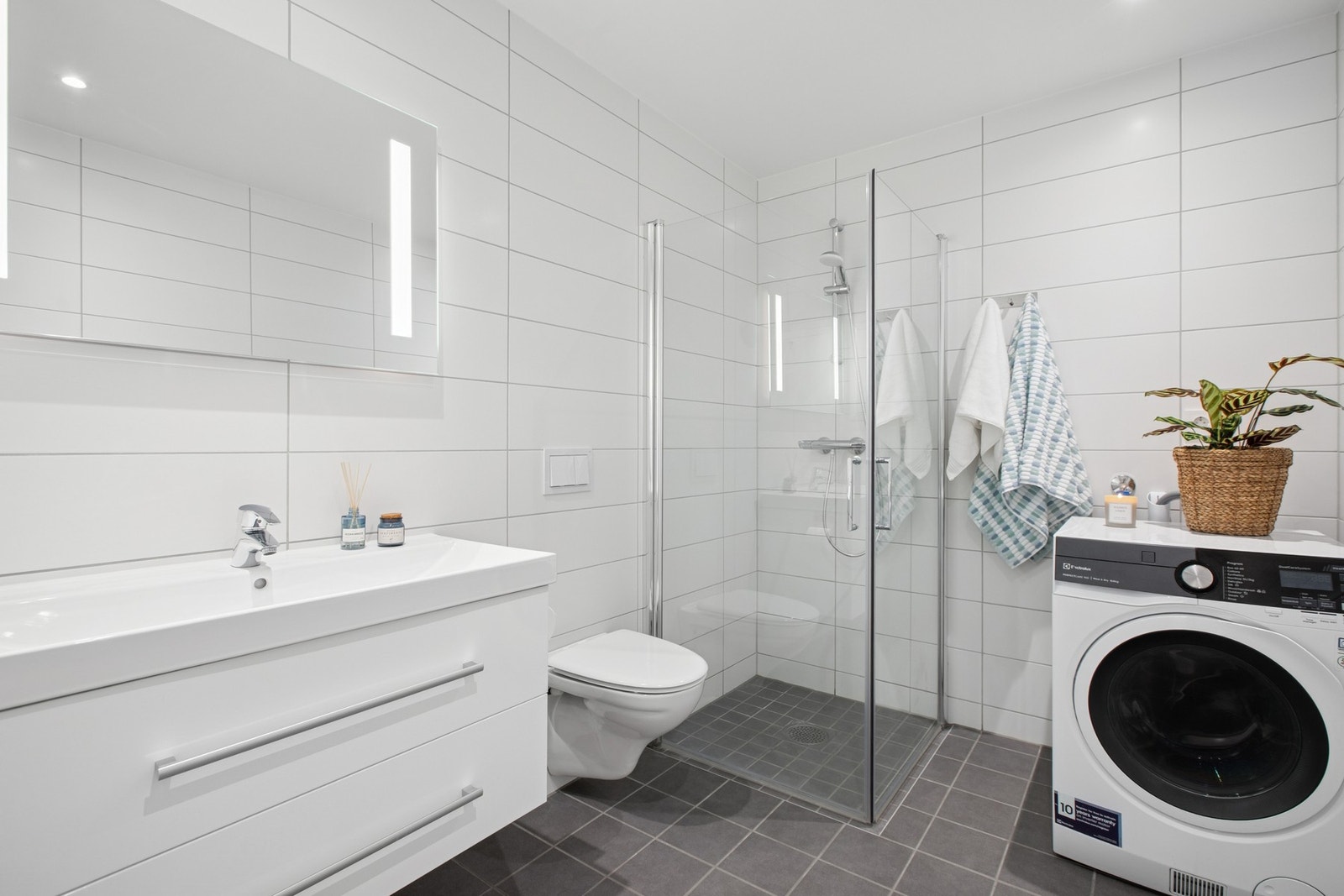 Badet i 1. etasje byr på en praktisk løsning med dusjhjørne, vegghengt toalett og opplegg til vaskemaskin. Lyse fliser og god belysning gir et rent og tidløst uttrykk. Galleribilde