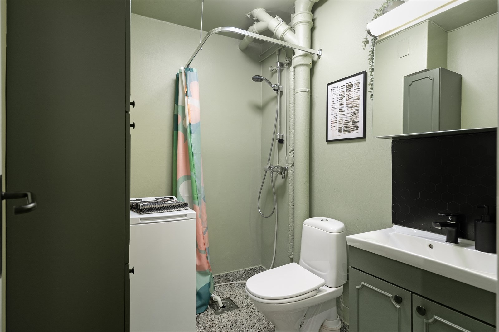 Badet er innredet med servant med skap, wc, åpen dusj og opplegg for vaskemaskin Galleribilde