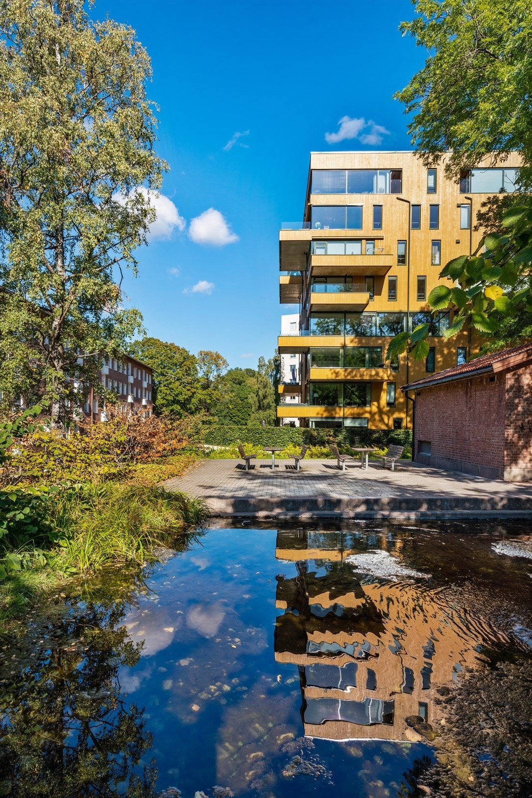 Sameiets tomt med dam, stor stenbelagt uteplass og plen, prydbusker, hekker og trær. Galleribilde