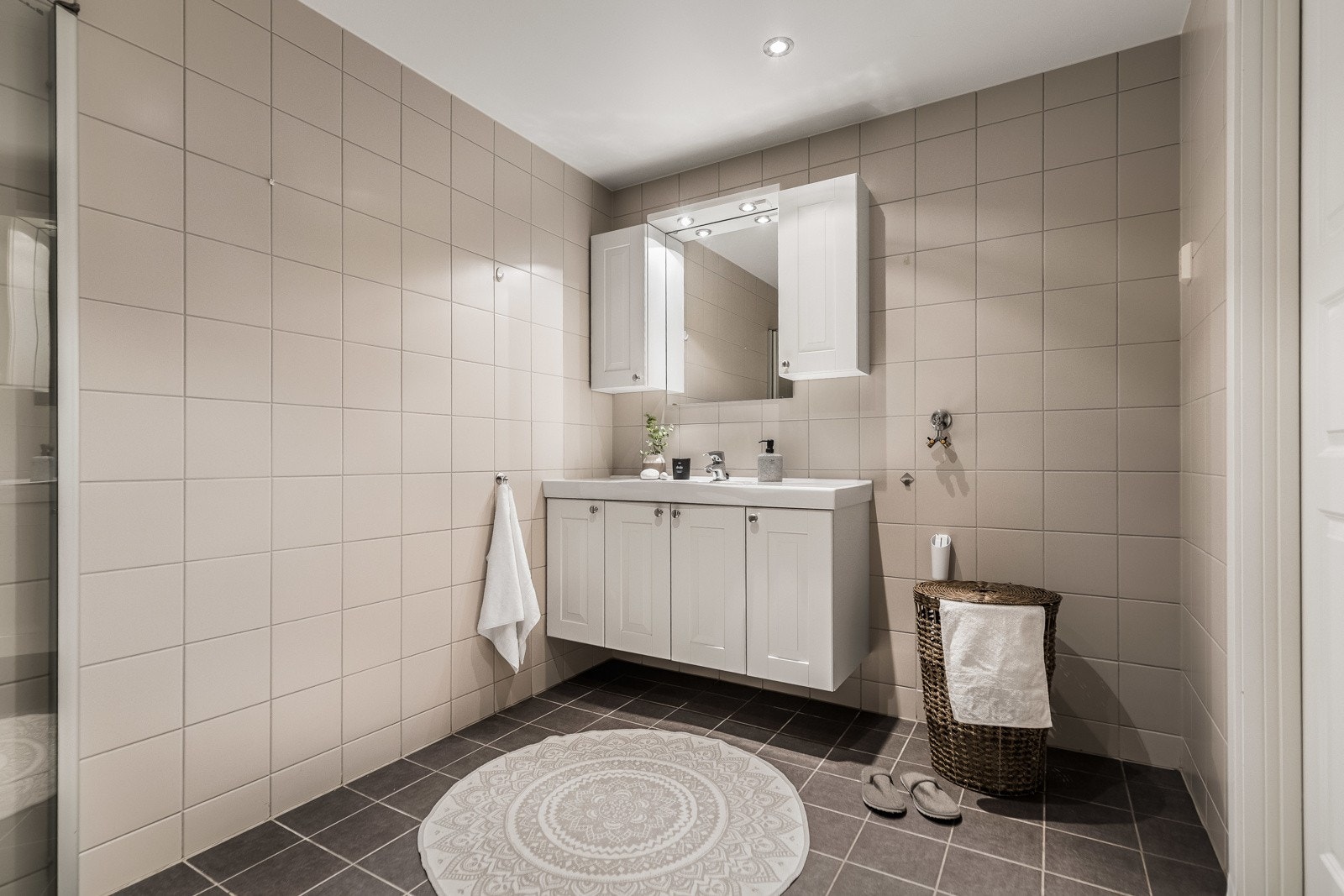 Badet har downlights i himling, mørke fliser på gulv med varmekabler og lyse fliser på vegg, samt opplegg for vaskemaskin/tørketrommel. Galleribilde