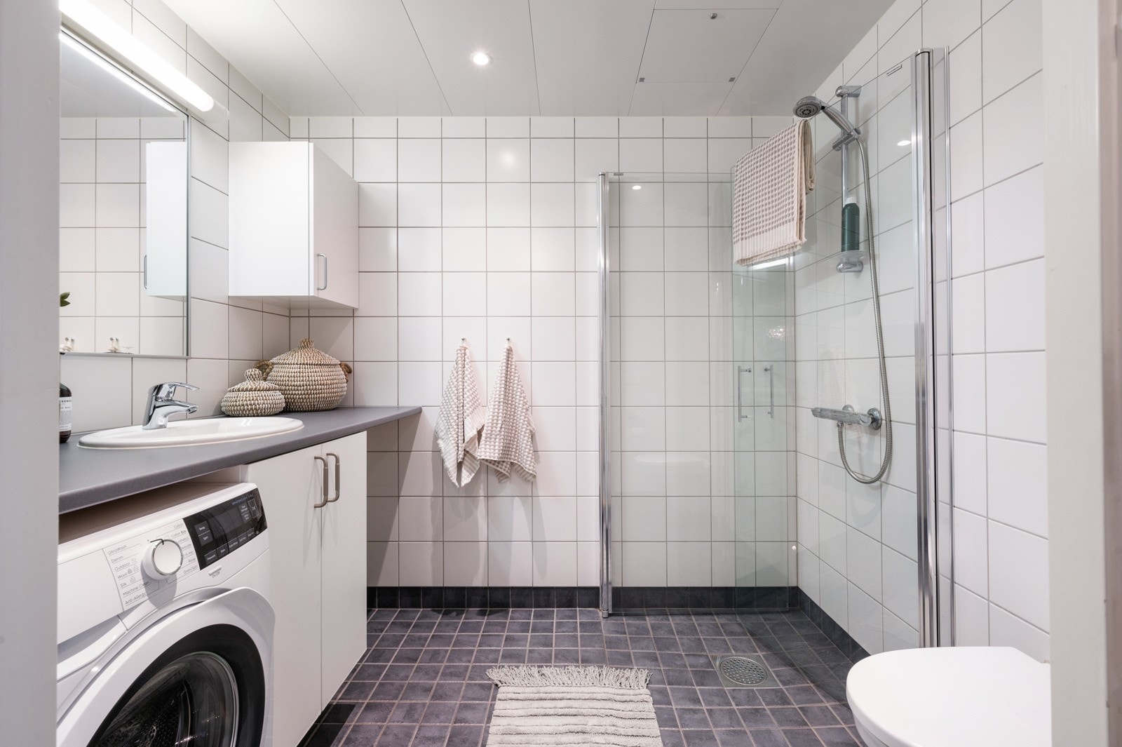 Moderne og stilrent baderom med flislagte overflater og innfelte downlights i taket. Praktisk løsning med vegghengt toalett og servant med oppbevaringsskap Galleribilde