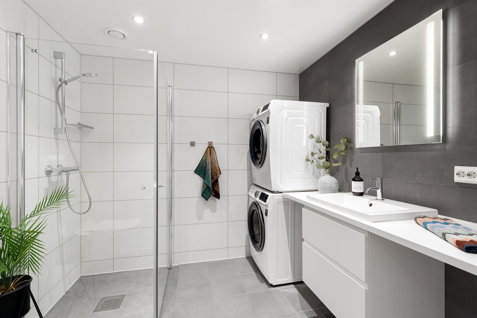 Tidløst og flislagt baderom med dushjørne, servant, speil over servant med lys , opplegg for vaskemaskin og vegghengt toalett Galleribilde