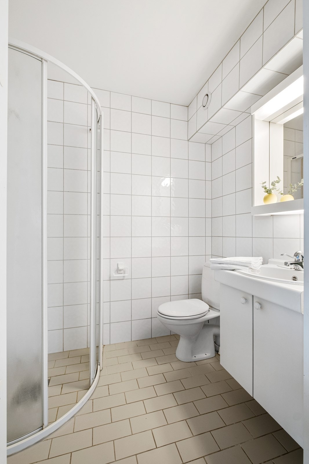 Fra entreen er det adkomst til en flislagt bad med varme i gulvet - badet er innredet med servant med skap under, speil over vasken, gulvmontert toalett og buet dusjhjørne Galleribilde