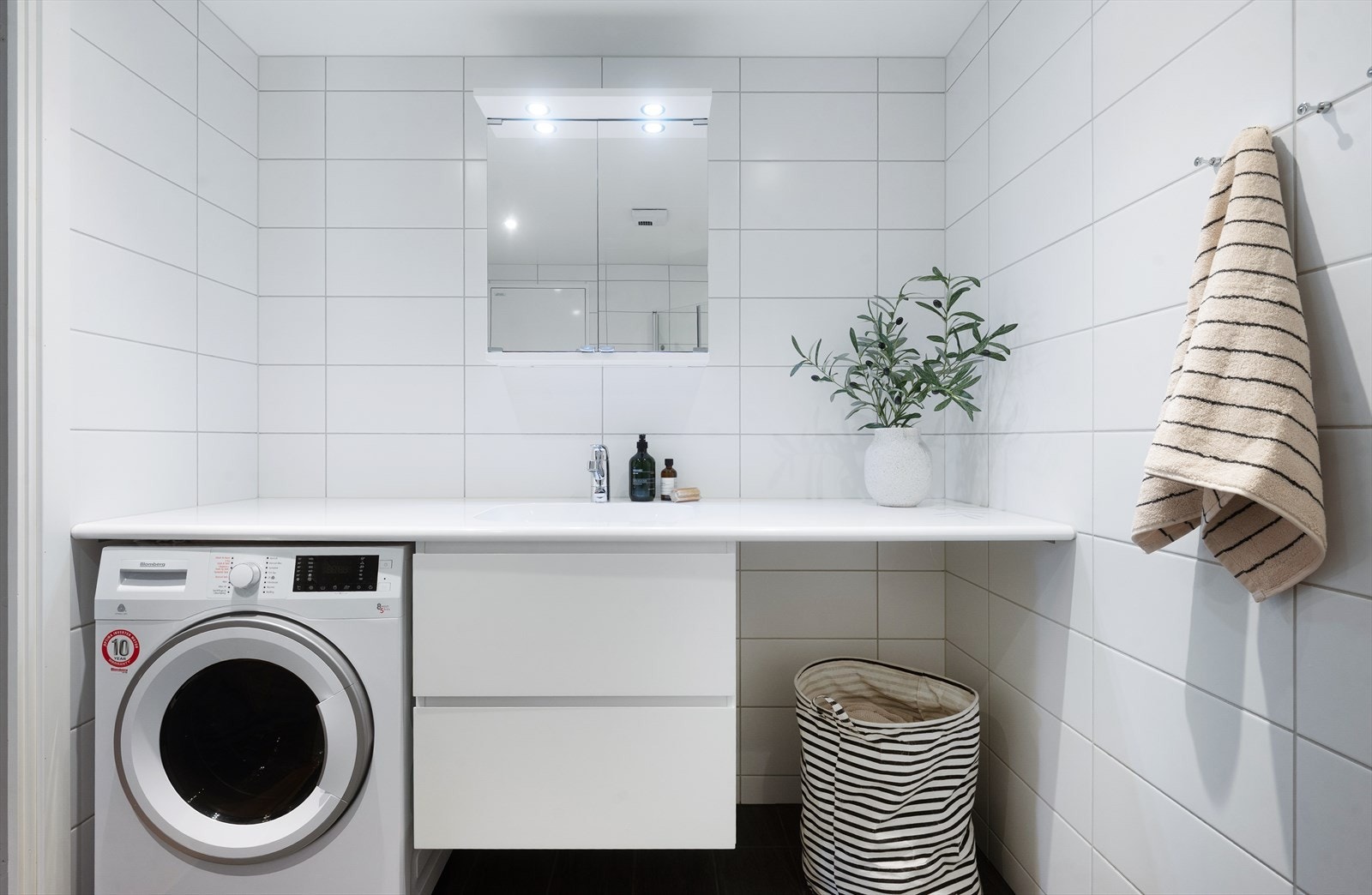 Baderomsinnredningen består av veggheng toalett, servant med underskuffer og benkeplate langs hel vegg, speilskap, og dusjhjørne med hånddusj og innfellbare glassdører. Under benkeplate er det plass til å sette inn vaskemaskin. Galleribilde