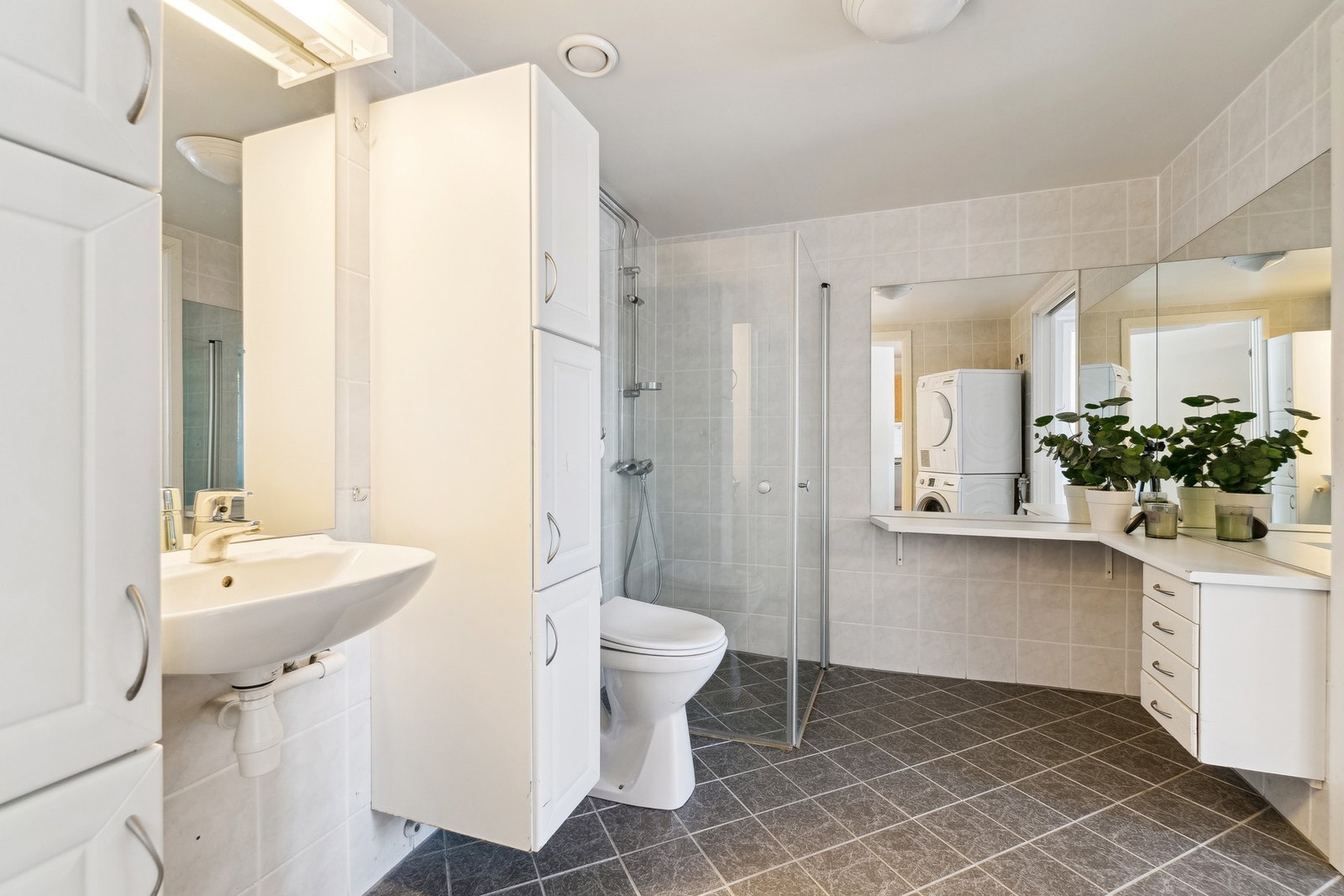 Badet har adkomst fra gangen, men også fra leilighetens hovedsoverom. Badet fremstår tidløst med en lys utførelse og mørke gulvfliser med varmekabler. Badet er utstyrt med servantskap, toalett og opplegg for vaskemaskin, samt en egen dusjsone i hjørnet so Galleribilde