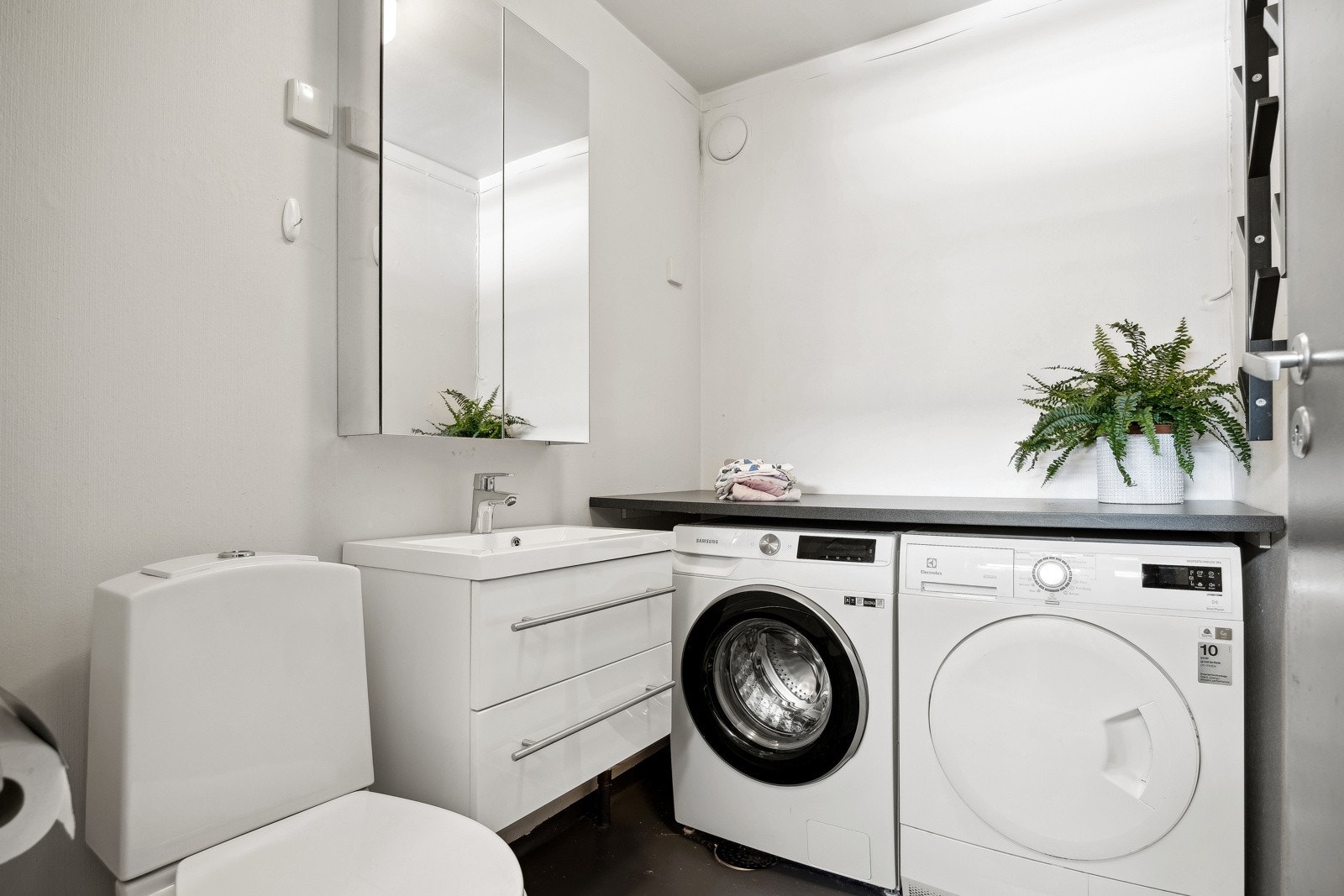Entréen fordeler seg videre til et praktisk vaskerom, innredet med toalett, nedfelt servant med skuffeseksjoner og speilskap. Det er god plass til både tørketrommel og vaskemaskin. Galleribilde