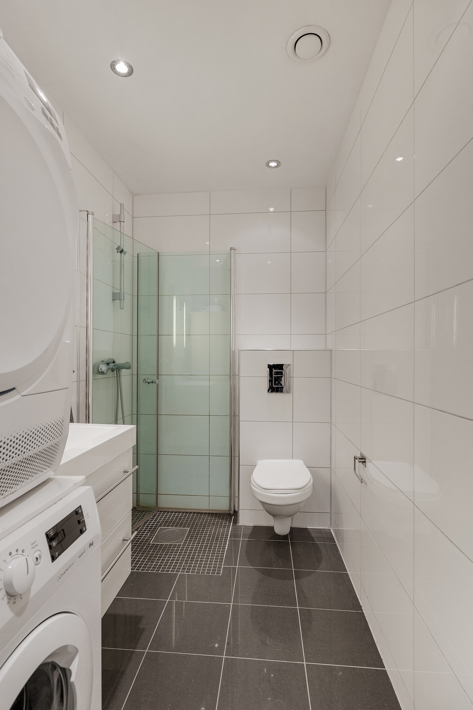 Badet har innfellbare dusjdører i glass, servantskap med skuffer, vegghengt toalett, opplegg for vaskemaskin, og plass til tørketrommel Galleribilde