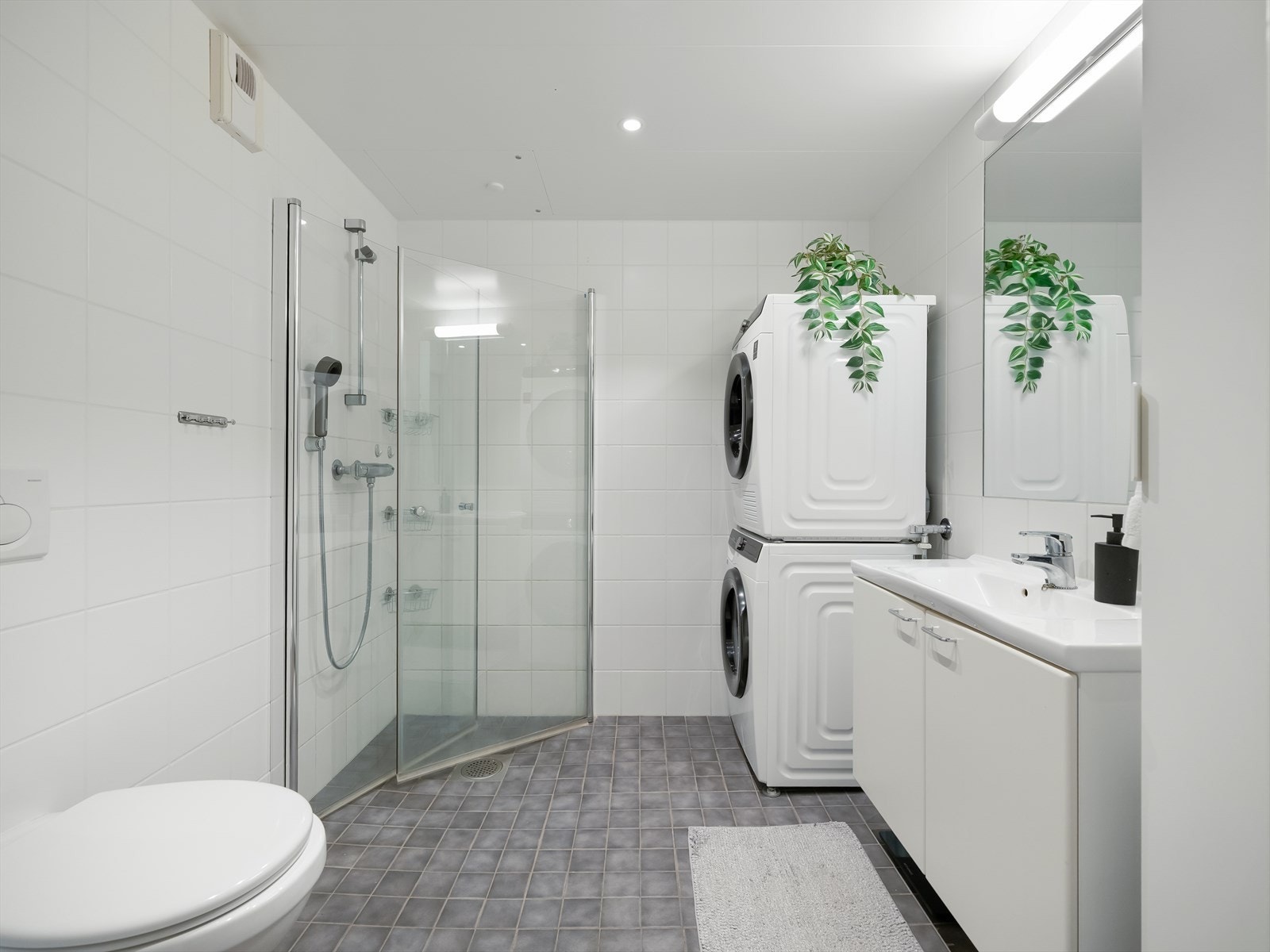 Pent flislagt bad med praktisk baderomsinnredning bestående av servant på underskap og speil med belysning over. Ellers installasjoner med vegghengt toalett, dusjhjørne og opplegg for vaskemaskin og tørketrommel. Galleribilde