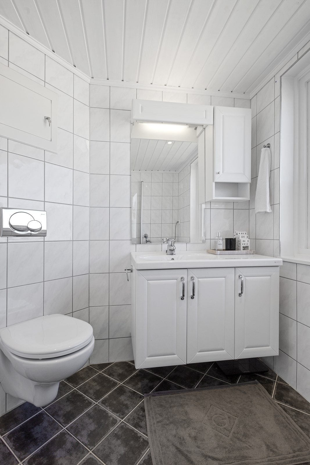 Det er innredet med hvit baderomsinnredning med heldekkende servant og med skap og hyller for å organisere toalettsaker og håndklær. Videre inneholder badet dusjhjørne og vegghengt toalett. Galleribilde