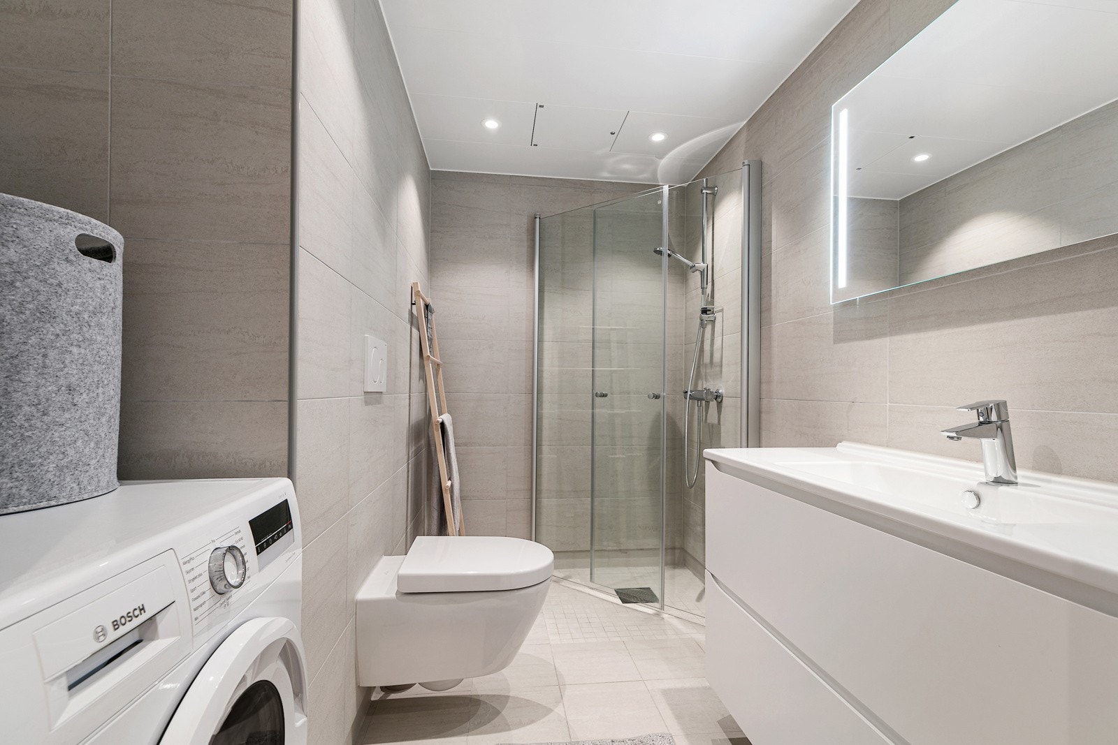 Et flislagt bad er delikat innredet med servant med skuffeseksjon, veggmontert toalett og et dusjhjørne med glassdører Galleribilde