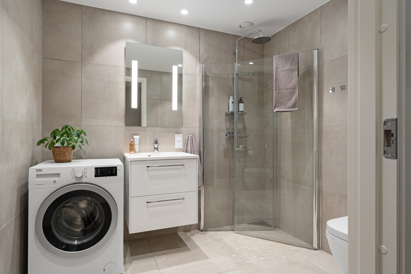 Lekkert bad med store beige fliser og utstyrt med praktisk vegghengt toalett, romslig dusjhjørne og plass for vaskemaskin. Galleribilde