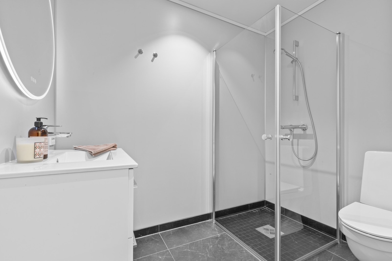 Pent baderom med flislagt gulv og varmekabler samt malte veggflater. Det er gulvstående toalett, dusjhjørne med glassdører og servant med innredning. Galleribilde
