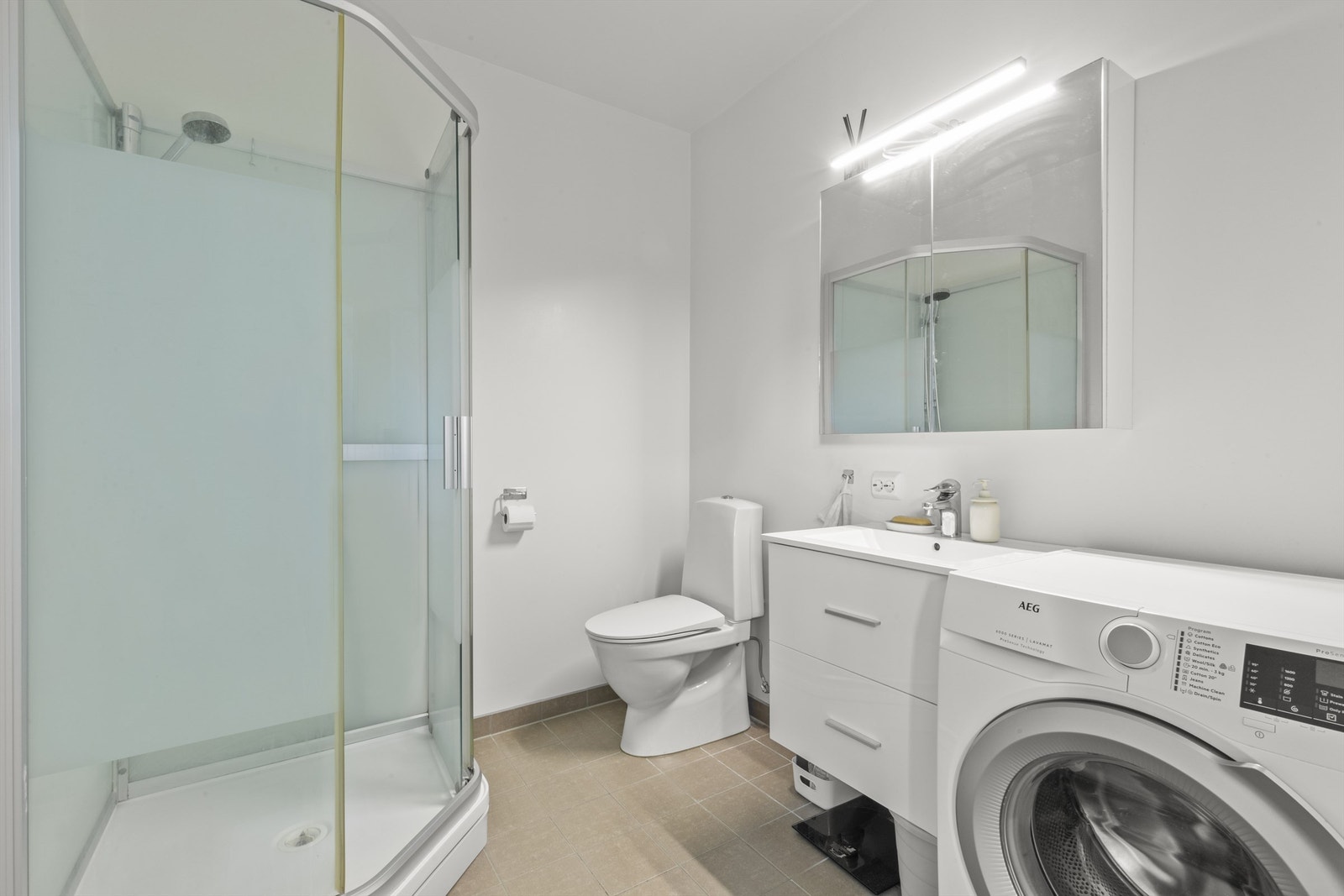 Baderom i hybelen med flislagt gulv og malte overflater for øvrig. Videre er det dusjkabinett, toalett, servant med innredning og vaskemaskin. Ellers speil over servant med innredning. Rommet varmes opp med varmekabler. Galleribilde