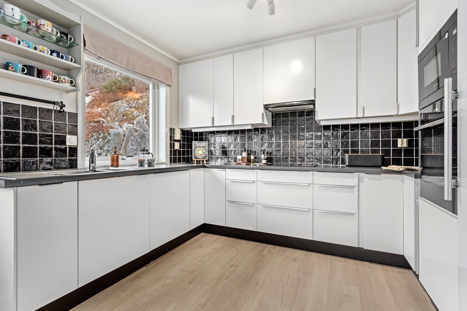 Kjøkkenet har kvit og glatt innreiing og benkeplate i laminat. På vegg mellom overskåp og benkeflate er det flis. Galleribilde