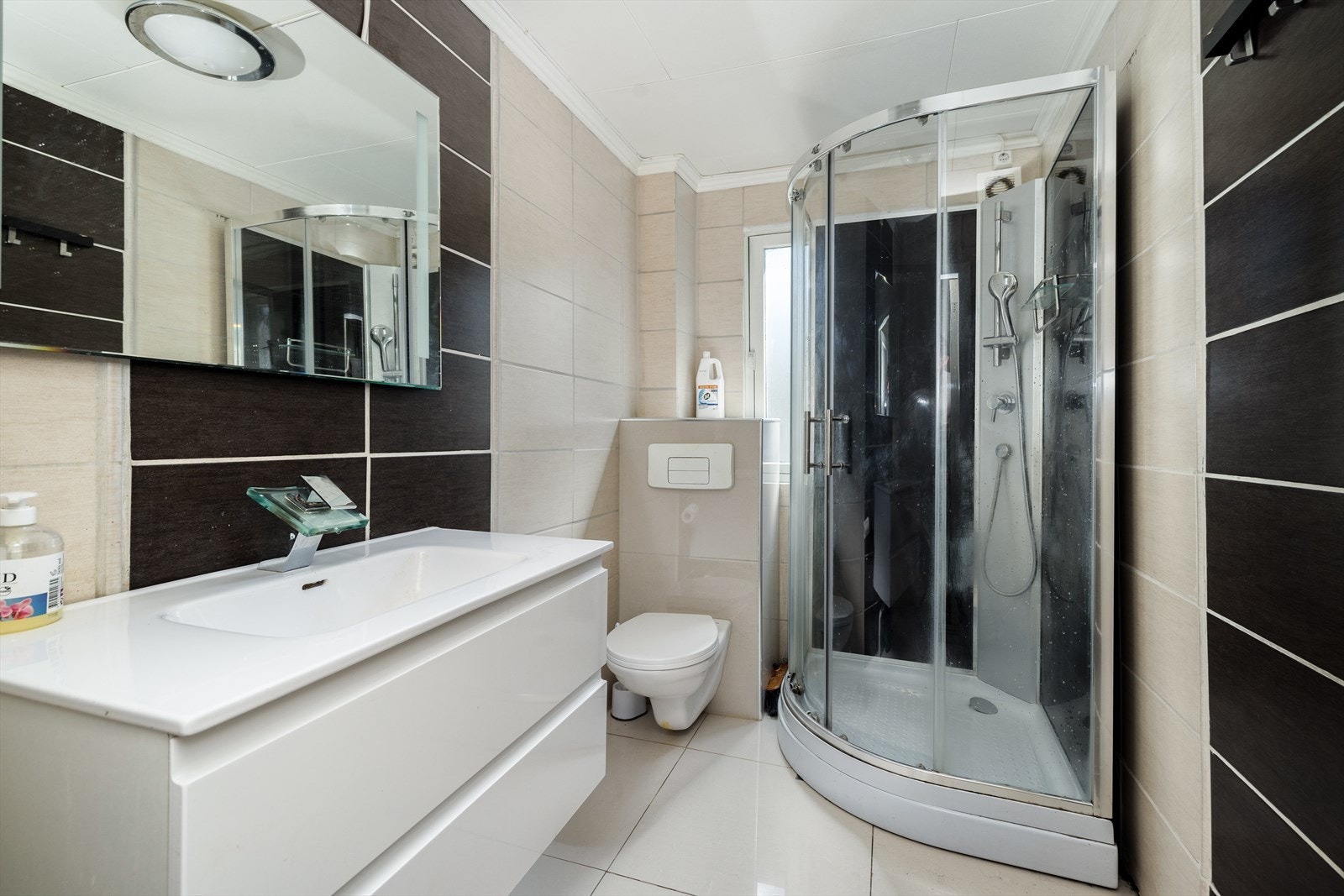 Flislagt bad med vegghengt toalett, dusjkabinett og servantinnredning med underskuffer og speil. Galleribilde