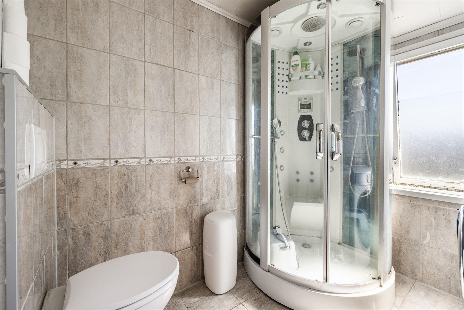 Flislagt bad som inneholder toalett, dusjkabinett og servantinnredning med underskap og speil. Galleribilde