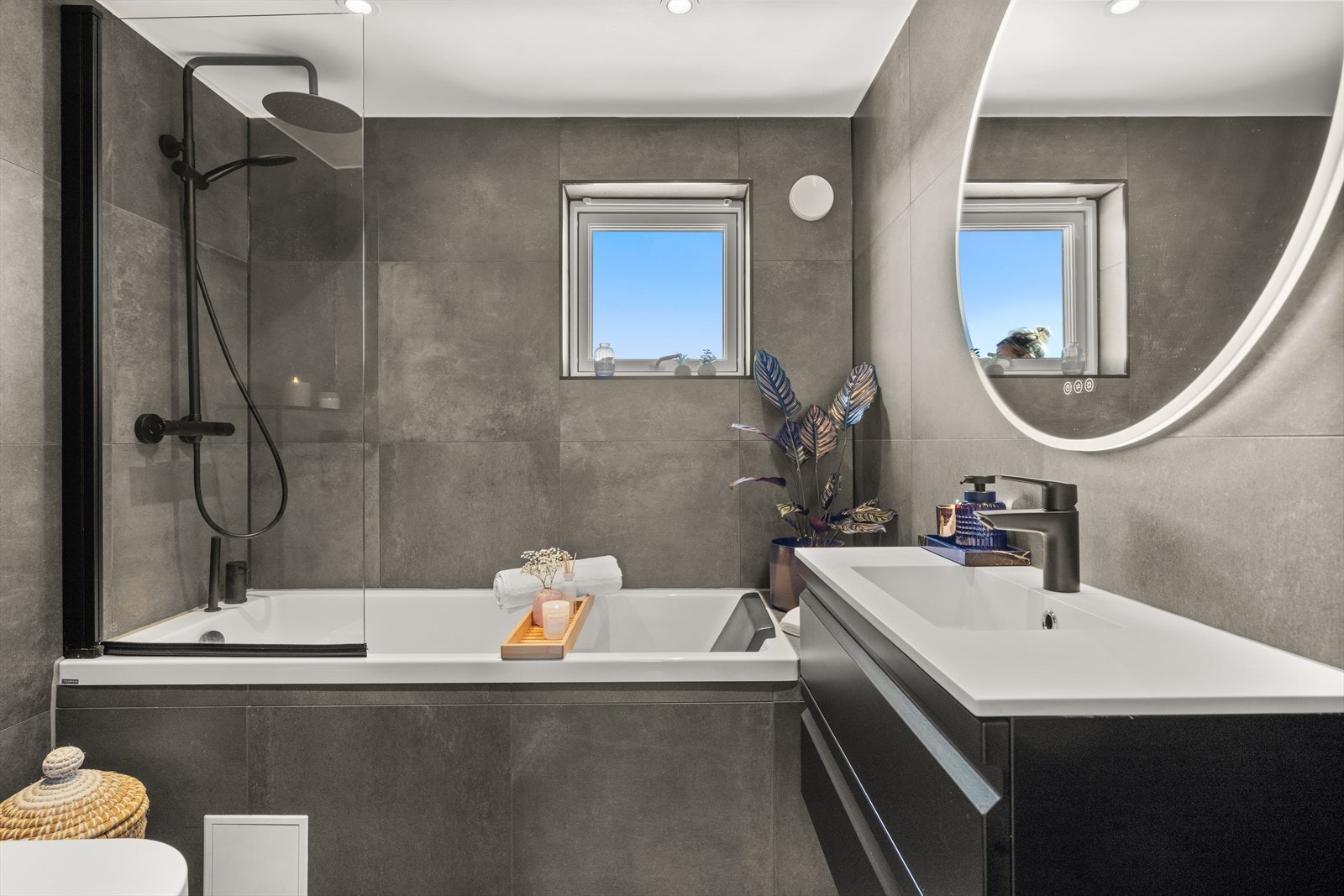 Grå fliser på gulv og vegger gir en god romfølelse. Badet er innredet med flislagt badekar, veggehengt toalett og servant med innredning. Galleribilde