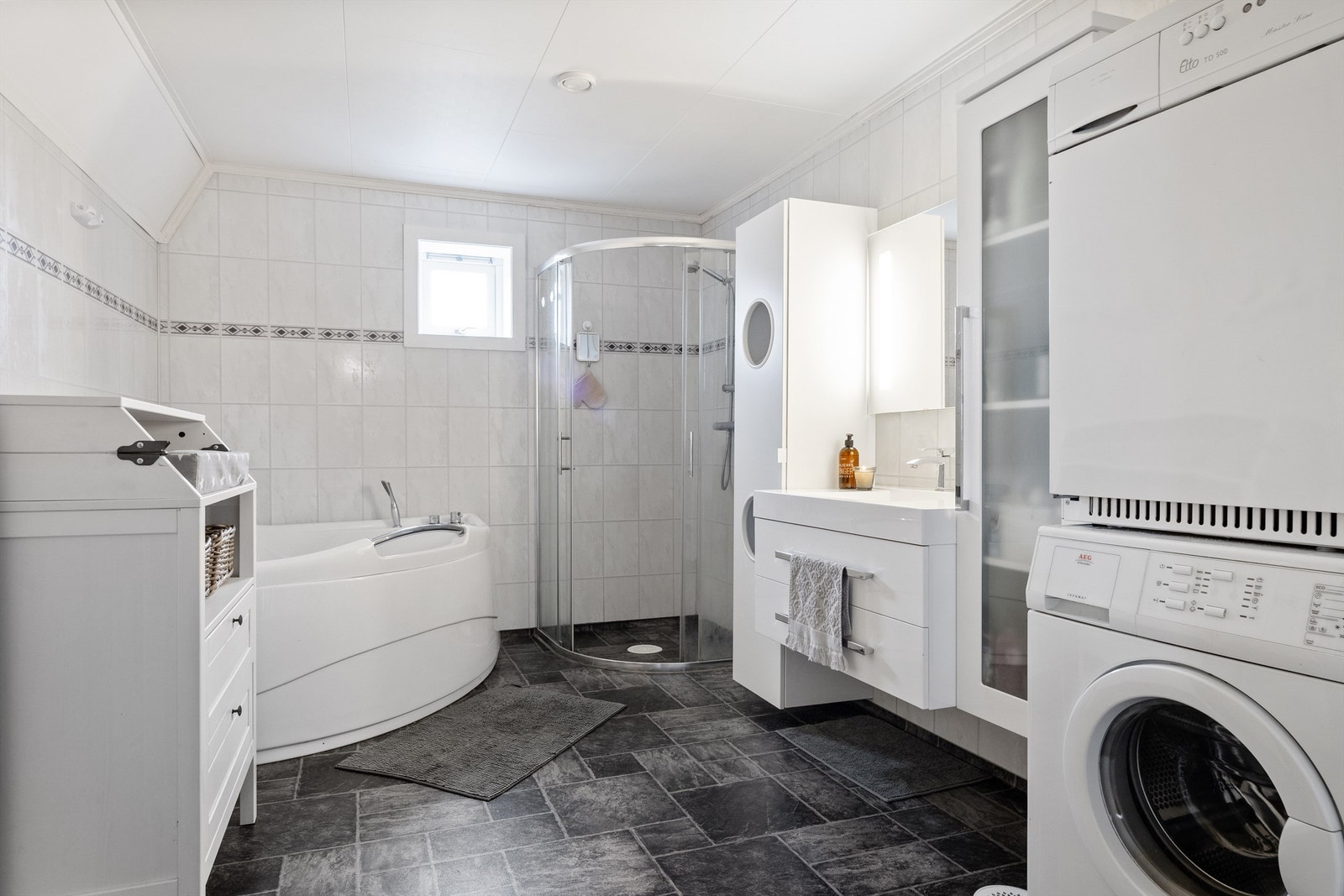 Stort og innholdsrik bad i 2.etasje. Badet er innredet med heldekkende servant med skuffeinnredning, to høyskap, hjørnebadekar, dusjhjørne med armatur på vegg og skyvedører, gulvmontert toalett og opplegg for vaskemaskin. Galleribilde