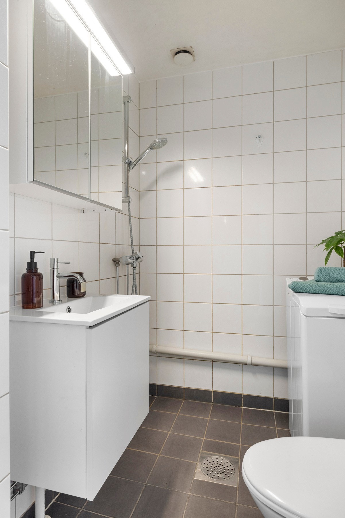 Lyst og flislagt baderom innredet med heldekkende servant med underskap (byttet i 2025), ettgreps blandebatteri og speilskap over servanten, gulvmontert toalett, dusjhjørne med Oras Oramix dusjbatteri fra 2023 samt opplegg for vaskemaskin. Galleribilde