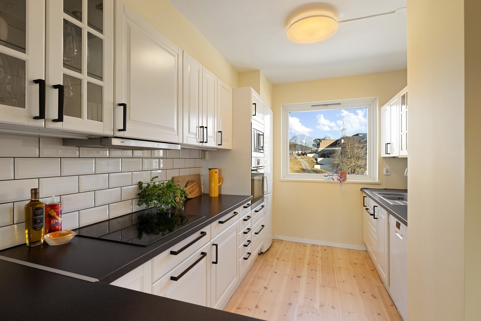 Lys innredning med hvite profilerte fronter, enkelte vitrinefronter, benkeplate i laminat og bistrofliser over kjøkkenbenk. Galleribilde