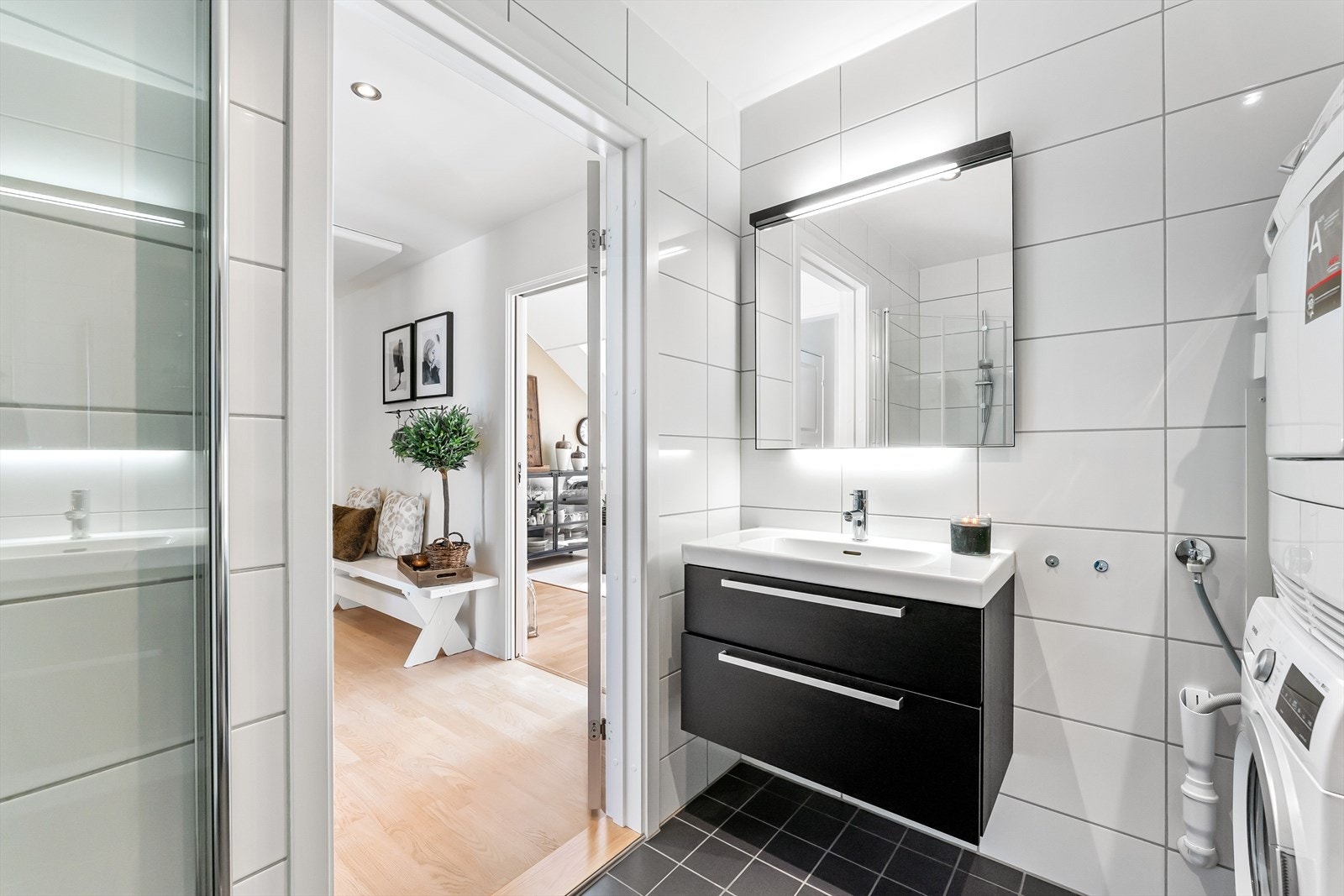 Videre består badet av vegghengt toalett, servant med underskuffer, speil og dusjhjørne med innfellbare glassdører Galleribilde