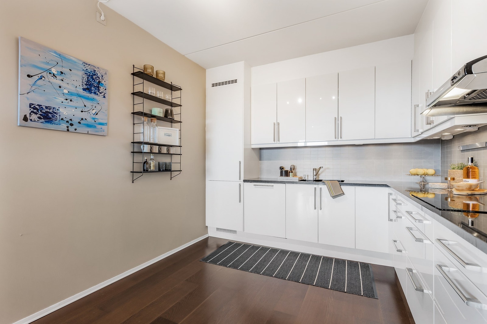 Overbelysning og stikkontakter over benk. Integrerte hvitevarer. Kjøleskap med fryser, oppvaskmaskin, stekeovn, mikrobølgeovn og platetopp. Vegghengt ventilator. Galleribilde