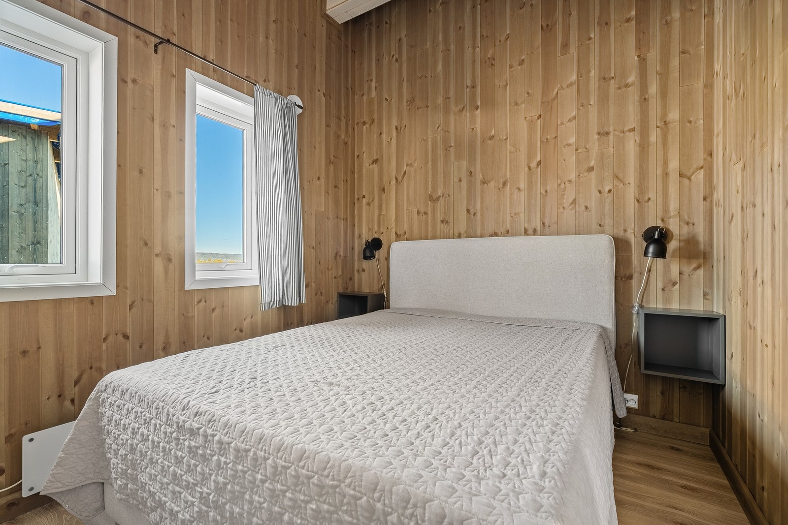 Soverom med panelvegger, to vinduer og en seng med grått sengeteppe. Galleribilde