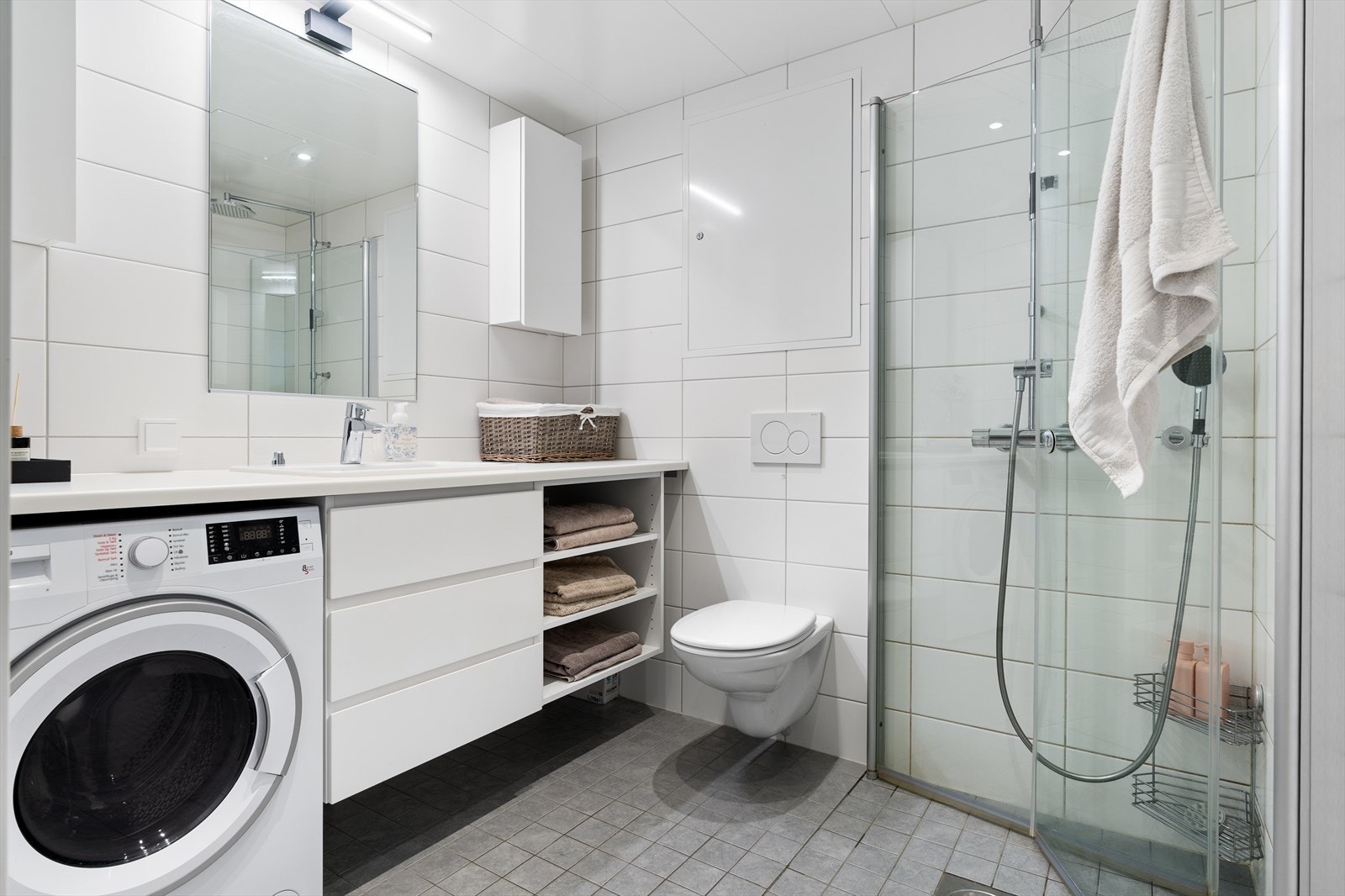 Lyst og lekkert bad med varmekabler i gulv og balansert ventilasjon. Praktisk baderomsinnredning med overbygd benkeplate og nisje for vaskemaskin. Galleribilde