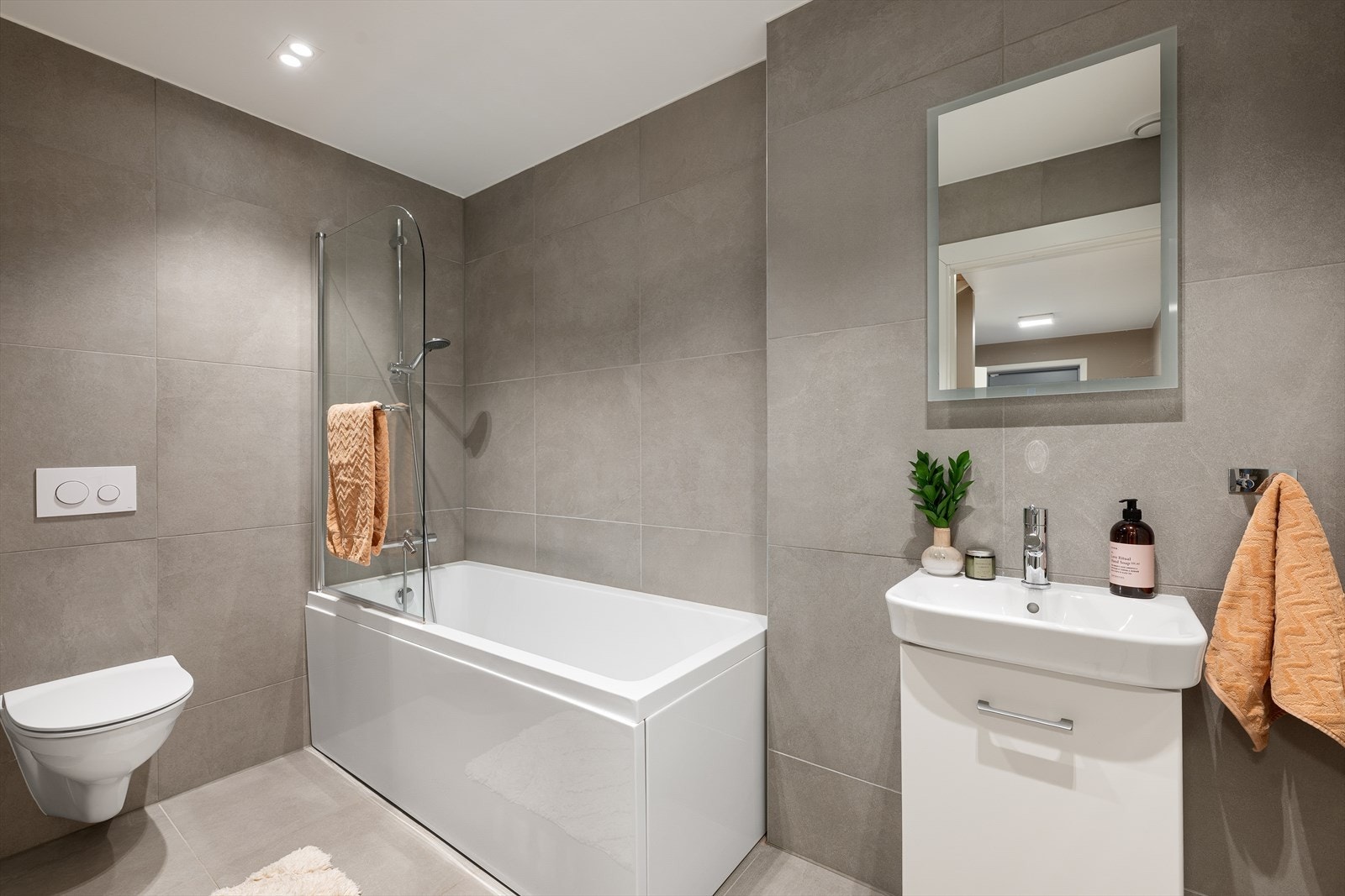 Flott flislagt baderom utstyrt med badekar og herdet glassdør, veggfestet klosett med innebygget sisterne og servant. Galleribilde