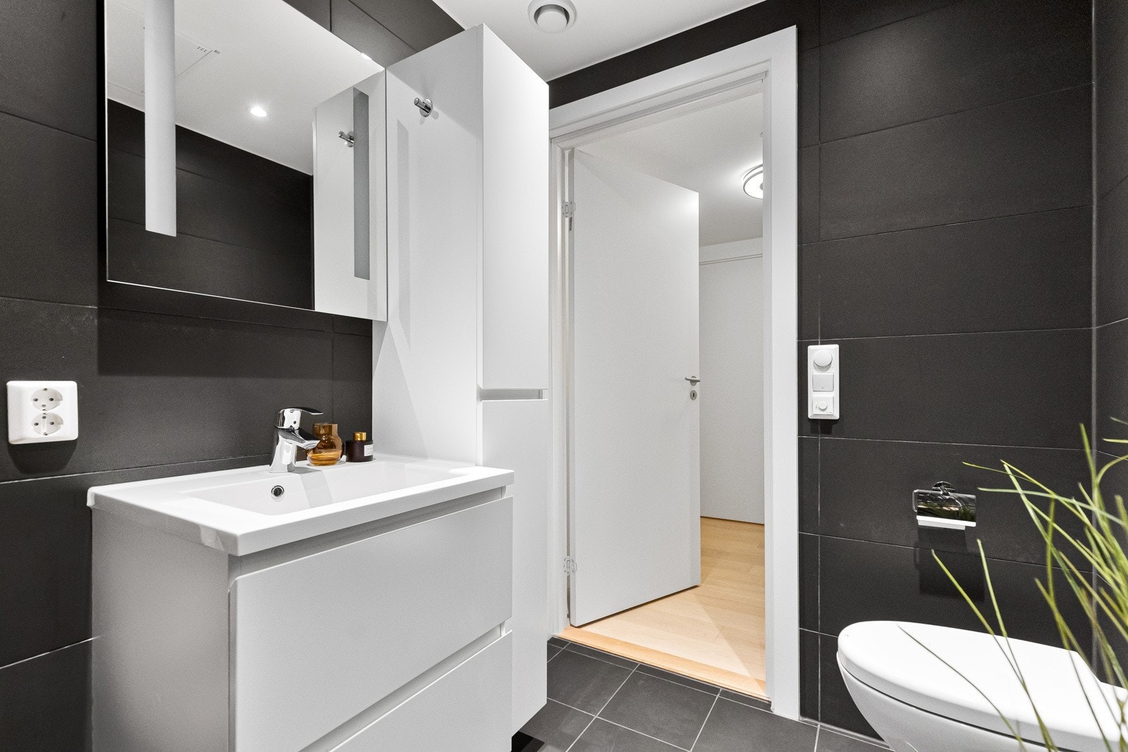 Badet er utført med moderne og stilrene fliser samt behagelig gulvvarme. Innredningen består av en nedfelt servant med godt med skapplass, vegghengt toalett og et dusjhjørne med glassdører. Galleribilde