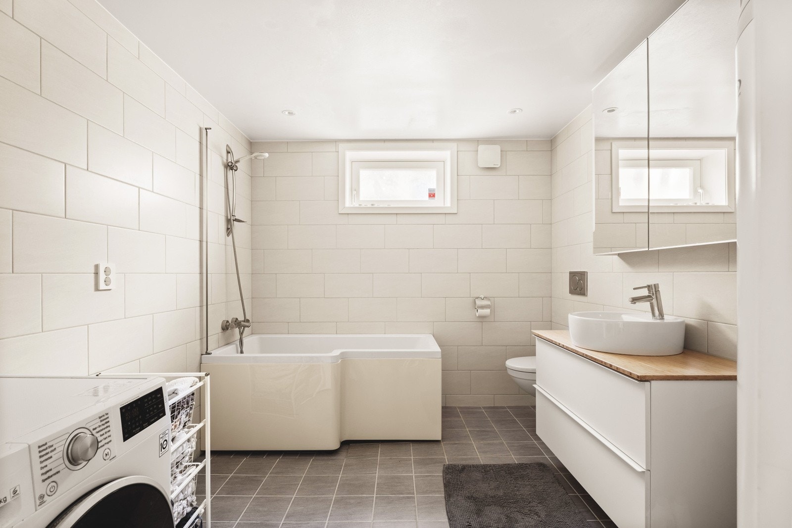 Hybelens bad har lyse flislagte vegger og gulv med varmekabler. Rommet er innredet med badekar, servant i innredning, vegghengt toalett og opplegg for vaskemaskin. Galleribilde