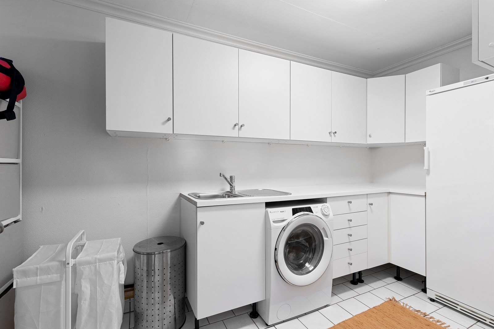 Praktisk og funksjonelt vaskerom med benkeplate og skapinnredning, Flislagt gulv og vegger med tapet. Rommet inneholder opplegg for vaskemaskin og vask/skyllekum. Galleribilde