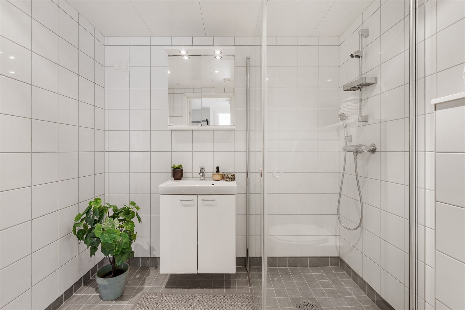 Innredning og garnityr på bad fremstår med: Veggmonterte dusjdører, servant med underskap, speilskap med lys over, opplegg for vaskemaskin og veggmontert wc. Galleribilde