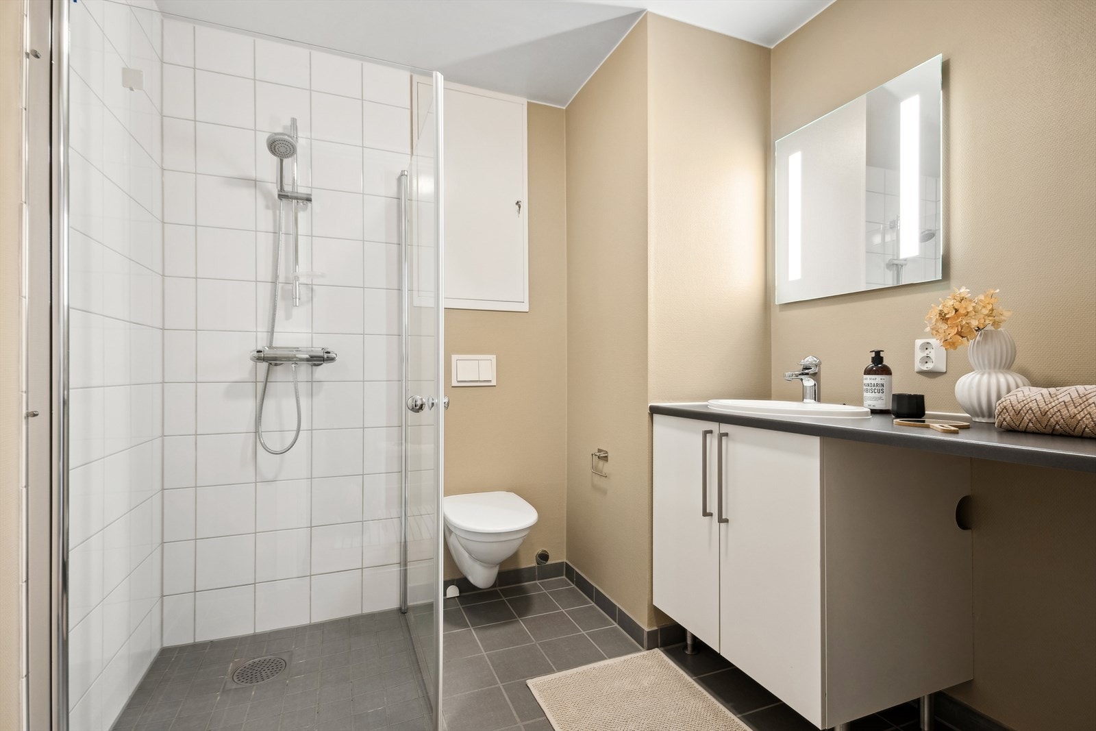 Badet er tidløst innredet med fliser på gulv og i dusjsone og malte flater. Badet er innredet med veggehengt toalett, dusjhjørne og servant med skaper. Galleribilde