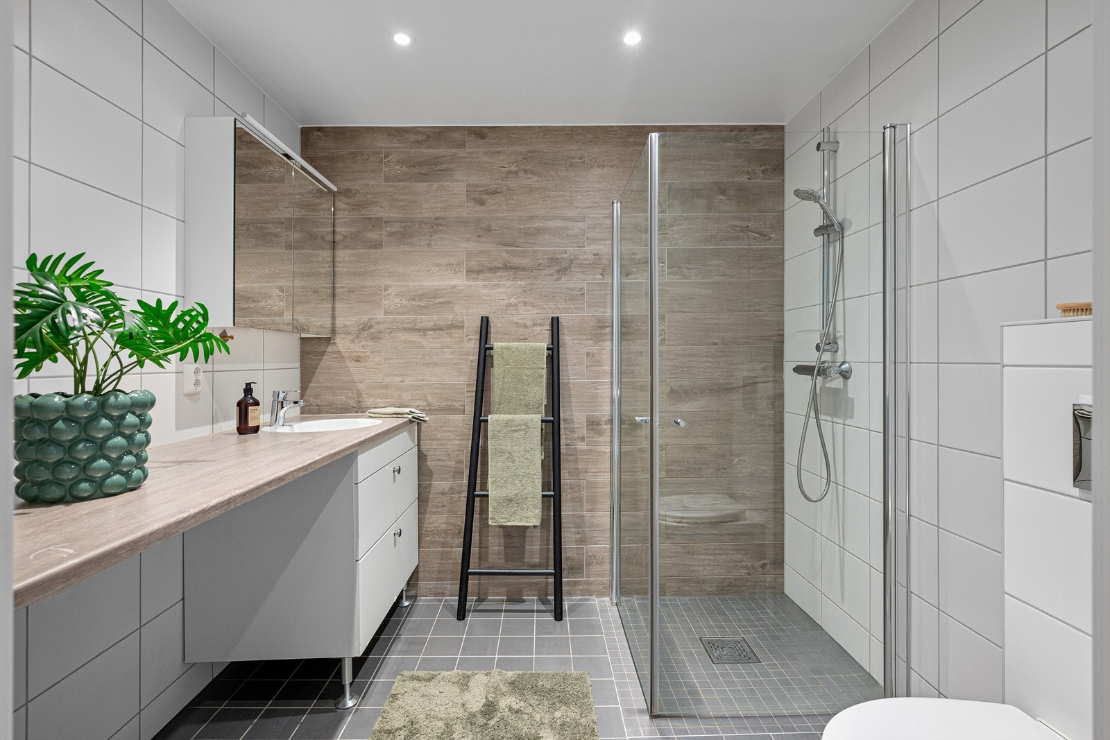 Lekkert flislagt bad med gulvvarme og downlights i nedsenket himling. Plass og opplegg for vaskemaskin og tørketrommel. Badet består av servant, skap, veggmontert toalett, dusjhjørne med nedfelt dusjssone og overskap med speilfront og lys Galleribilde